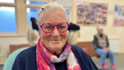 BBC An elderly women is smiling at the camera. She has pink framed round glasses on and a pink and purple scarf. She has her hair in pigtails with a black scrunchies. 