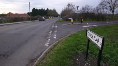 A rural road bordered by grassy verges and leafless trees. A black car is driving away in the distance. On the right side of the image, a white street sign reads “TODWICK ROAD S26” near a junction with traffic islands and bollards.