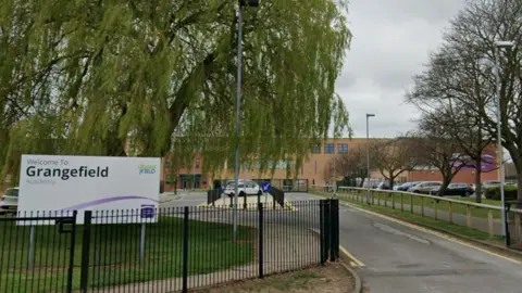 Google The exterior of the school from behind it's black gates. A purple and white sign on the field reads: "Welcome to Grangefield Academy". Down the driveway to the building is a car park. The orange brick building is long and three-storeys tall.