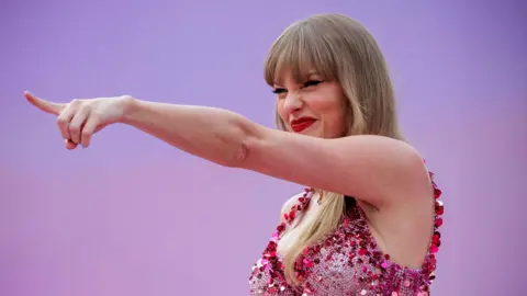 PA Taylor Swift points into the distance with a grin and her eyes squinted beneath her blonde fringe. She wears a sparkly pink fitted bodice and red lipstick. The background is pinkish-purple gradient and matches her outfit.