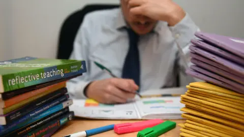 PA Media A teacher sits at a desk with their head in their hand, appearing stressed while marking work. Open exercise books and colourful highlighters are on the table. Piles of folders and a stack of teaching guides, including “Reflective Teaching,” surround them.