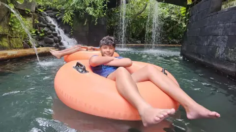 Supplied A young boy, with his arms crossed, floating on a inflatable orange ring in a pool with greenery and a waterfall in the background 