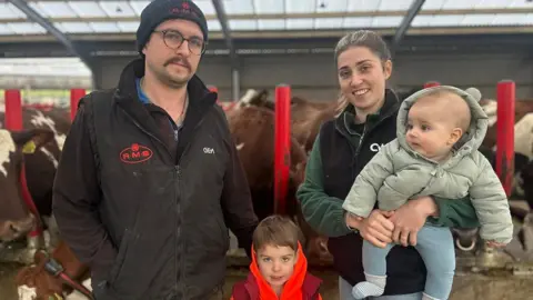 BBC News Farmers Adam Johnstone, wearing a black gilet and black hat and Lucy Johnstone, wearing a green fleece and black gilet, standing in front of a cow shed, with Lucy holding her 5-month-old son and their 4-year-old son standing in between them 