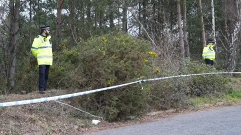 Shaun Whitmore/BBC Police tape surrounding a wooded area next to a road with two officers standing on scene guard.