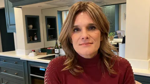 BBC Ellen looks at the camera with a small smile. She is in a kitchen in a house, with the cabinets and work surface visible behind her. She has short dark blonde hair, which is loosely curled, and wears a red jumper with diamante sequins on the shoulders. 