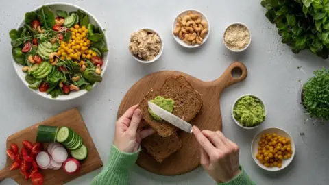 Natalia Gdovskaia/Getty Images A photo from above of a table with bowls of chickpeas, mashed avocado, nuts, seeds and salads. In the middle of the table, a pair of hands are spreading avocado on slice brown bread