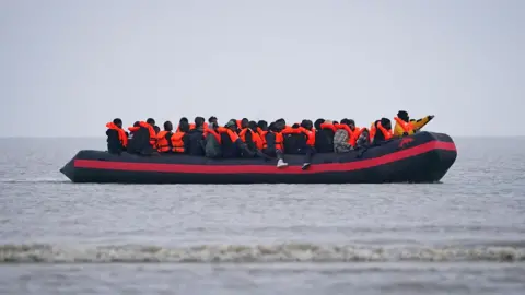 PA People thought to be migrants onboard a small boat in Gravelines, France, on 7 November 2025. They are wearing orange life vests and the boat is black and orange.