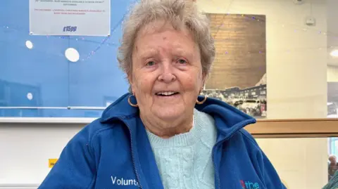 Groundwork Sheila Underwood with short grey hair wearing a blue fleece, pale blue jumper and gold hoop earrings in a community centre. She is sitting at a desk with a noticeboard behind her. She is smiling.