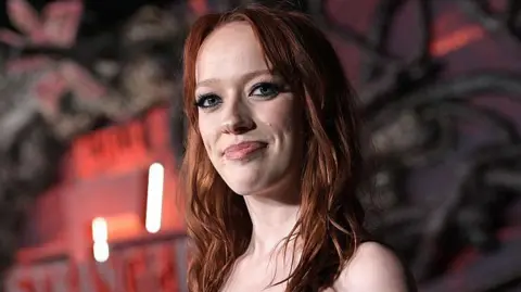 Charley Gallay/Getty Images Marybeth is pictured at a film premier. There is a black and red background with strobe lighting. She is smiling at the camera and has ginger hair. 