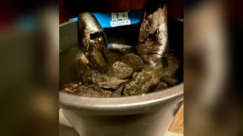 Emma Lamport A black bucket filled with water, with black leather Victorian shoes floating in it and a sign behind which reads Beach Academy