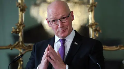Getty Images A bald man with glasses and wearing a black jacket, white shirt and purple tie has his hands pressed together as if in prayer
