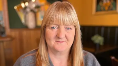 BBC Joan has long blond hair and is wearing a jumper