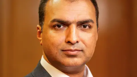 Manchester City Council A promotional headshot image of Councillor Shahbaz Sarwar, with short hair and wearing a grey suit and white shirt.