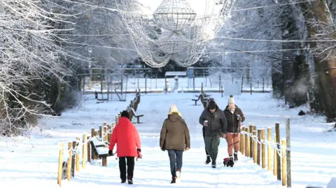 Pacemaker Four people wearing winter clothing walk along the footpath at Antrim Castle Gardens. The ground is covered in snow, and the trees are white. Above those walking, a large chandelier is hanging.